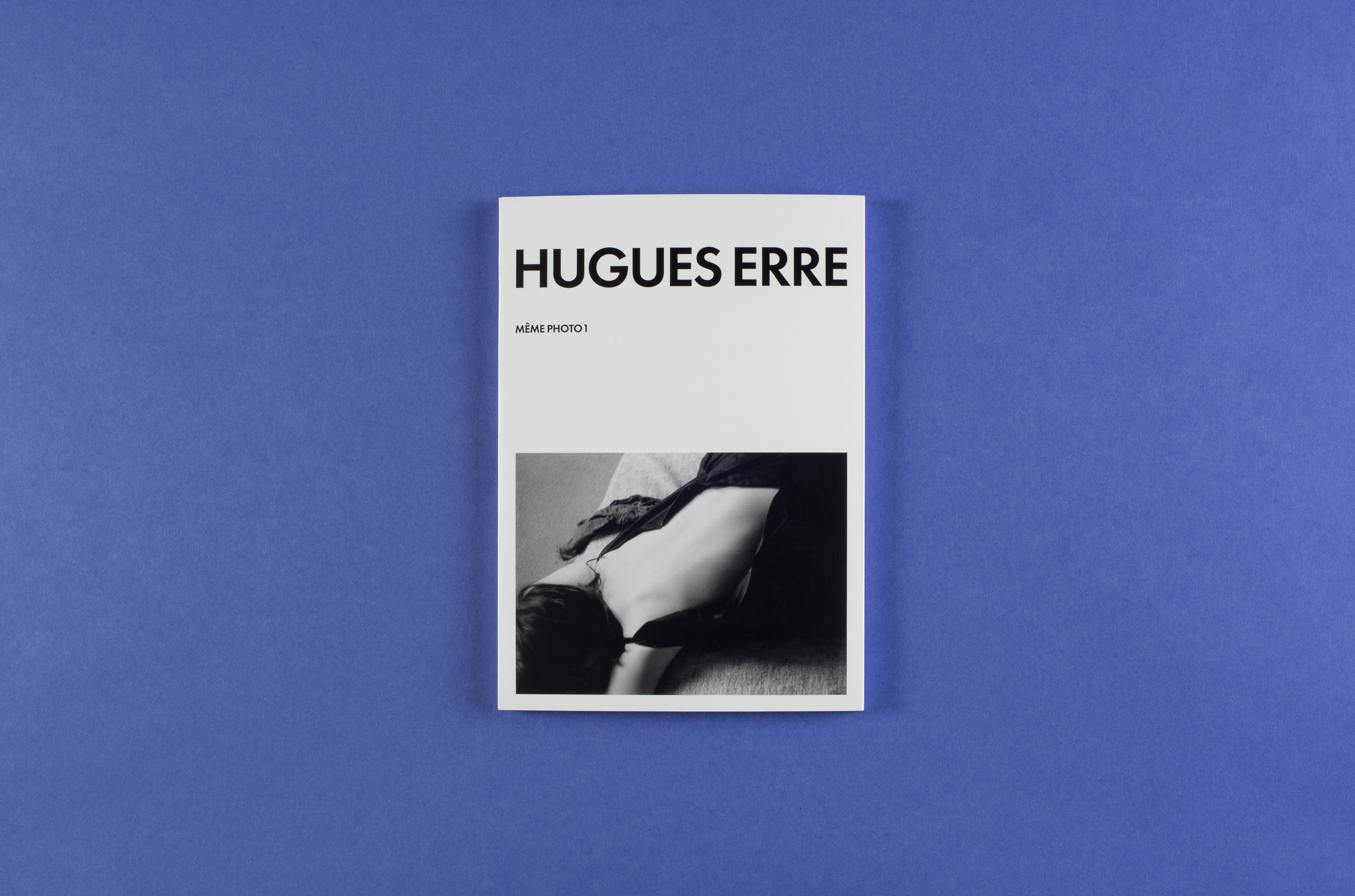 Photo of a white photo book on a blue background. The cover of the book shows the name of the photographer ‘Hugues Erre’, the name of the book series ‘Même Photo 1’ and a black and white photograph of a woman’s back.