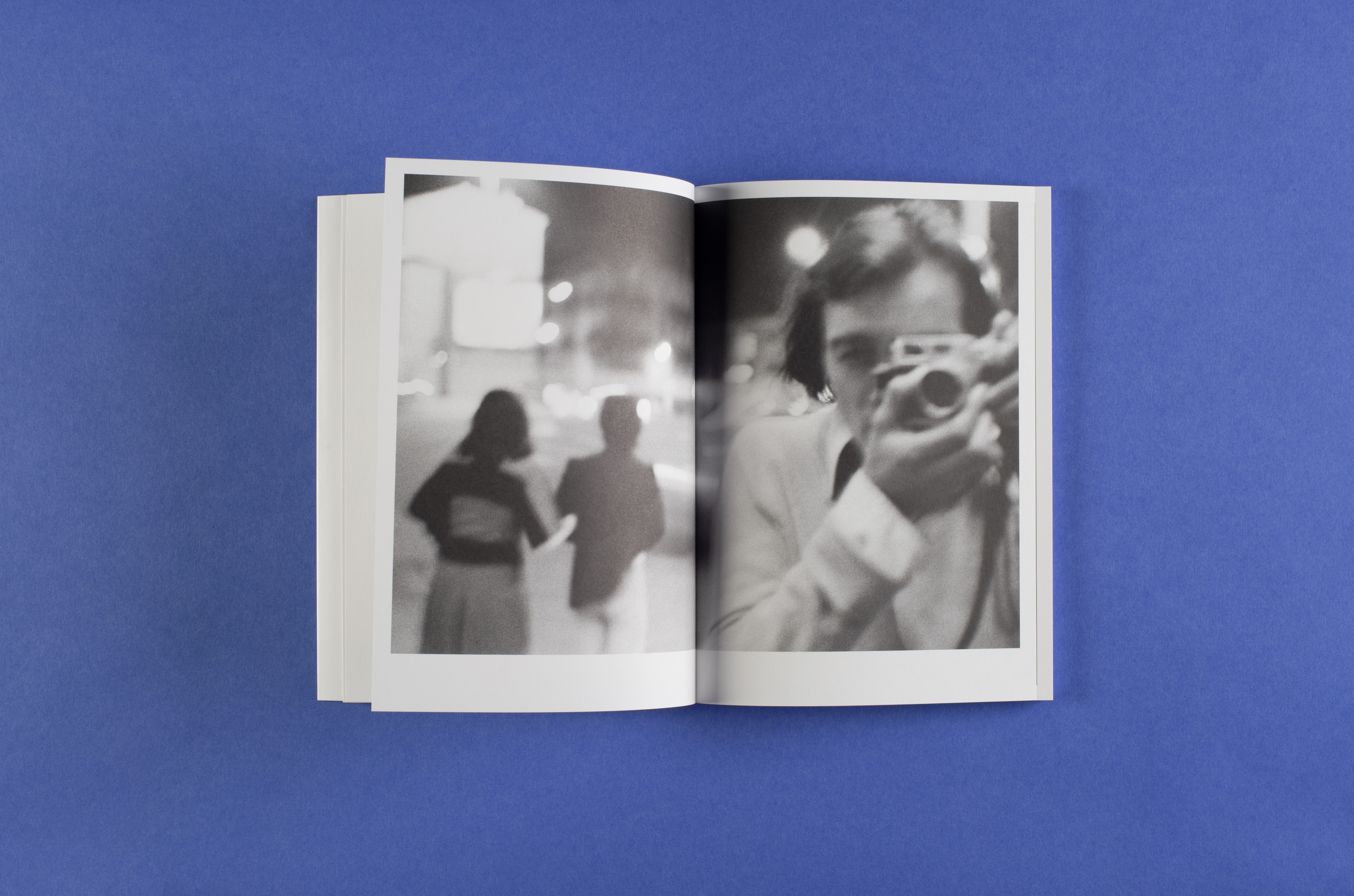 Photo of the open photo book in landscape format. One can see a photograph that runs across both pages. The picture shows the photographer Hugues Erre, photographing himself in a mirror or another reflective surface and two women from behind. The photo was taken at night, as it shows the lights of a city illuminated at night.