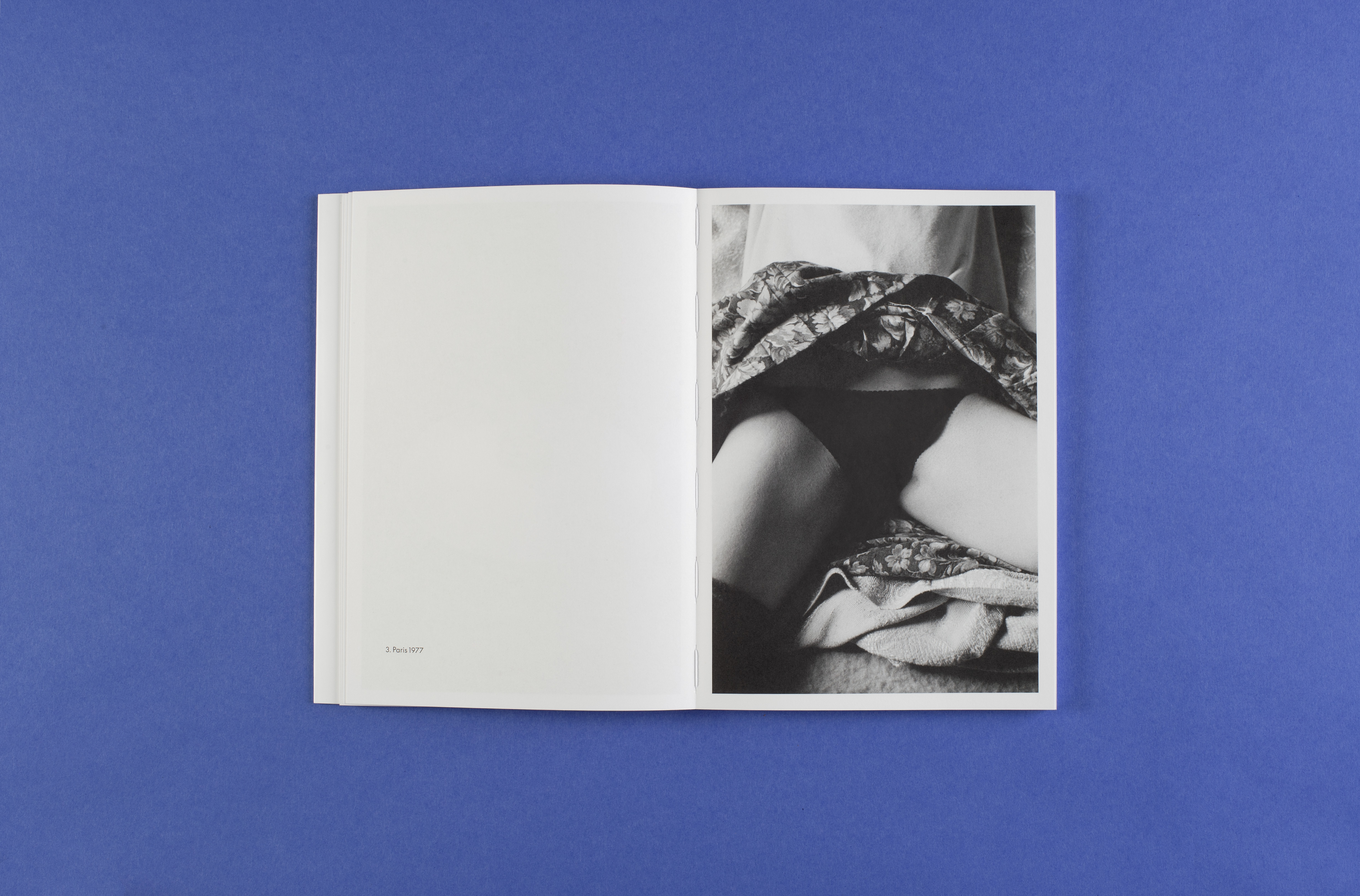 Photo of the open white book on a blue background. It shows a black and white photograph of a female abdomen in black pants and a floral skirt pulled up. The title of the picture is: Paris 1977.