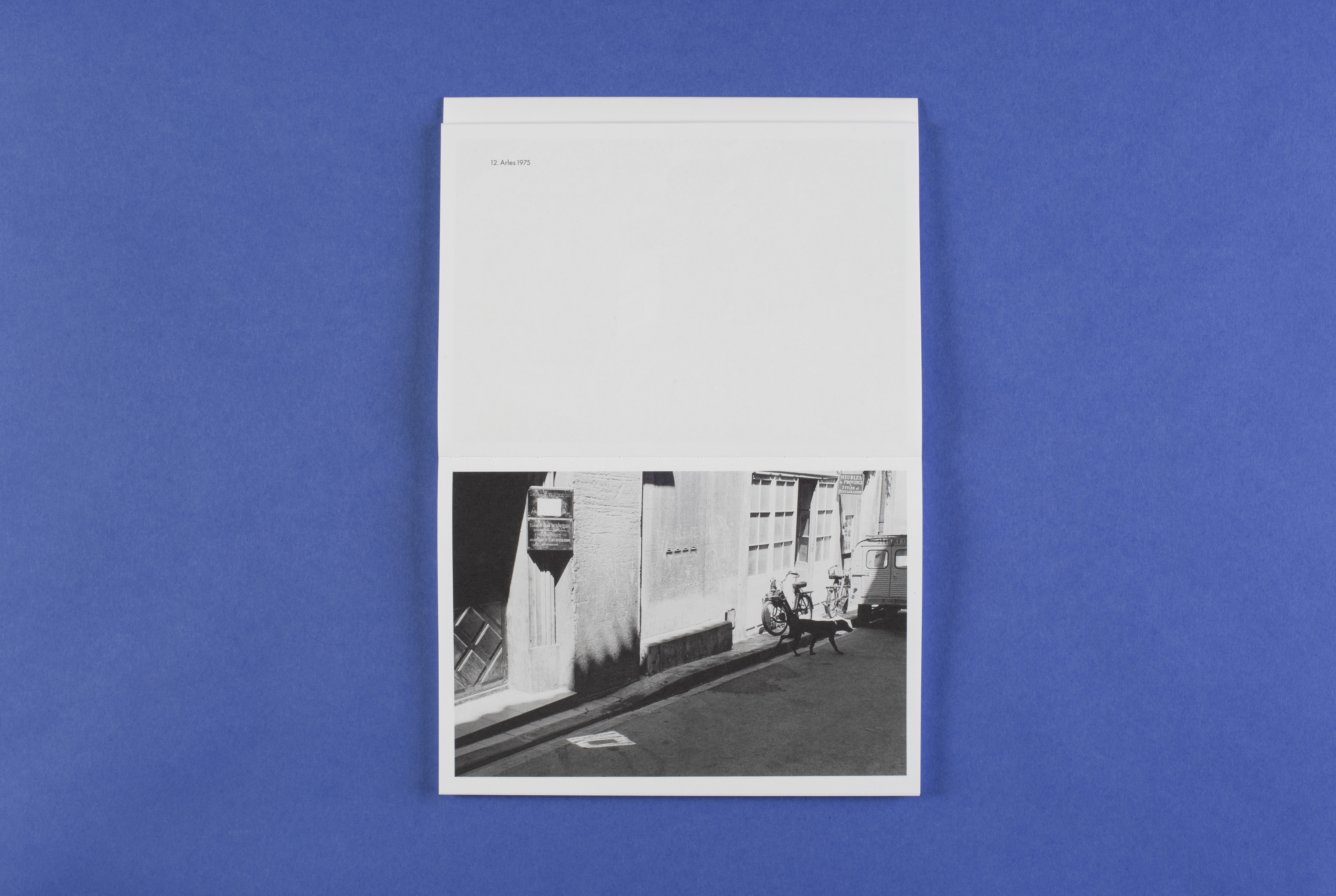 Photo of the open photo book in portrait format. The photo shows a house wall glistening in the sunlight and a part of a street and a part of a car. A black dog walks through the scene. The title of the picture is: Arles 1975.