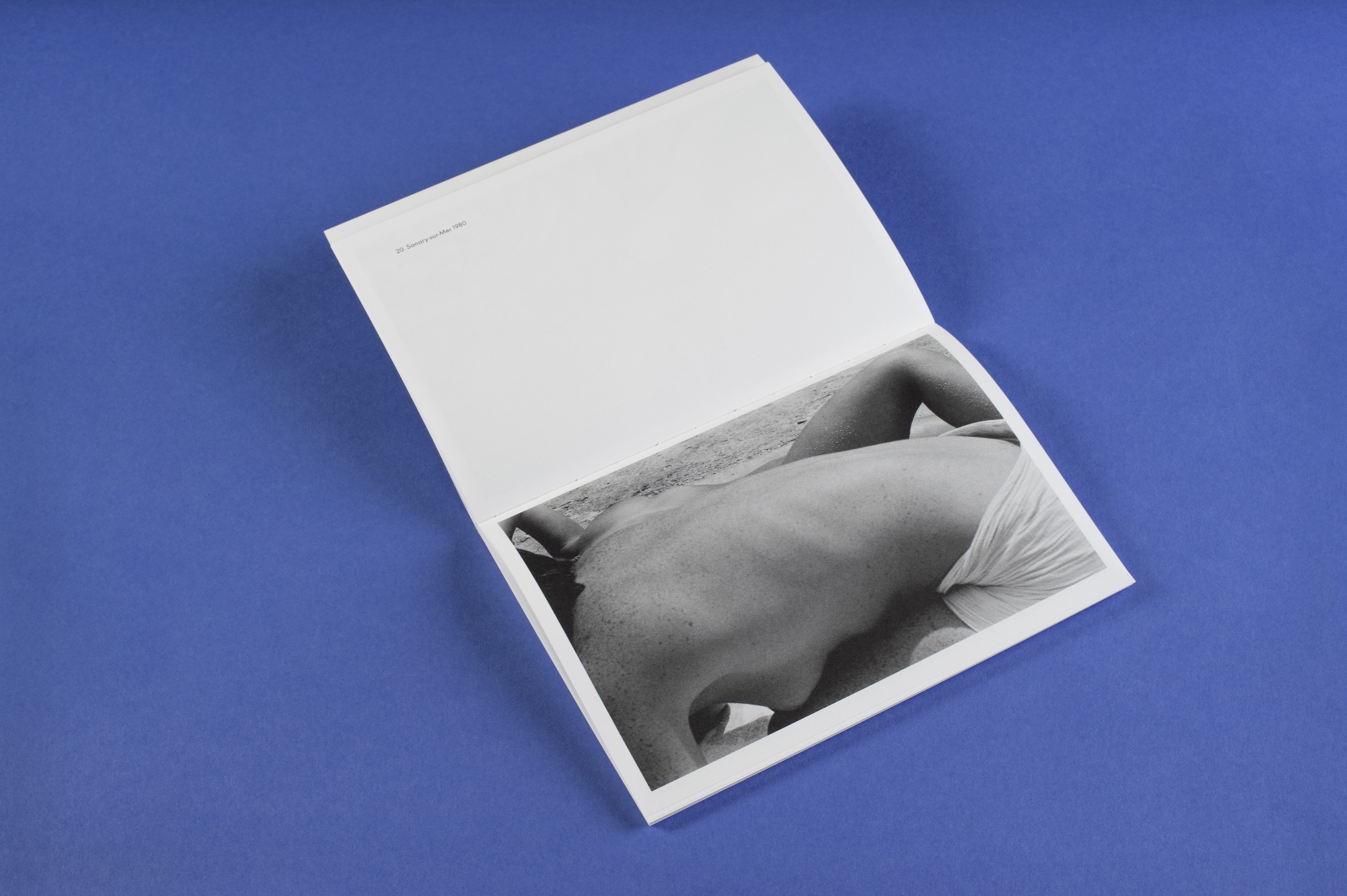 Photo of the open photo book in landscape format taken from diagonally above. The photo shows a naked female torso from the side. Behind it is a part of a female upper body lying on her back. The picture was taken on a beach. The title of the picture is: Sanary-sur-Mer 1980.